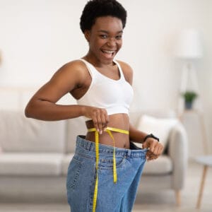 Programme Neuro-minceur - Black Lady Measuring Waist Wearing Oversized Jeans After Slimming Indoors
